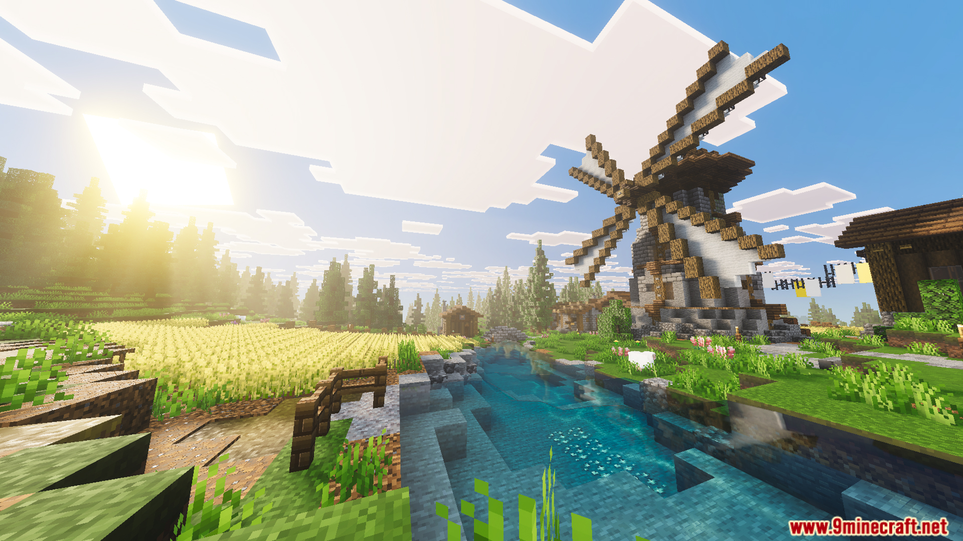 A tall windmill stands next to a flowing river and wheat fields