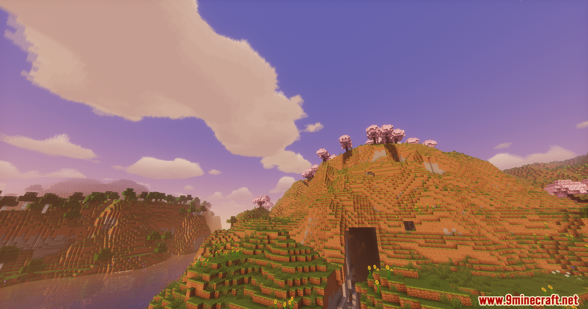 Bright sunlight shines on green hills and pink trees near a cave entrance