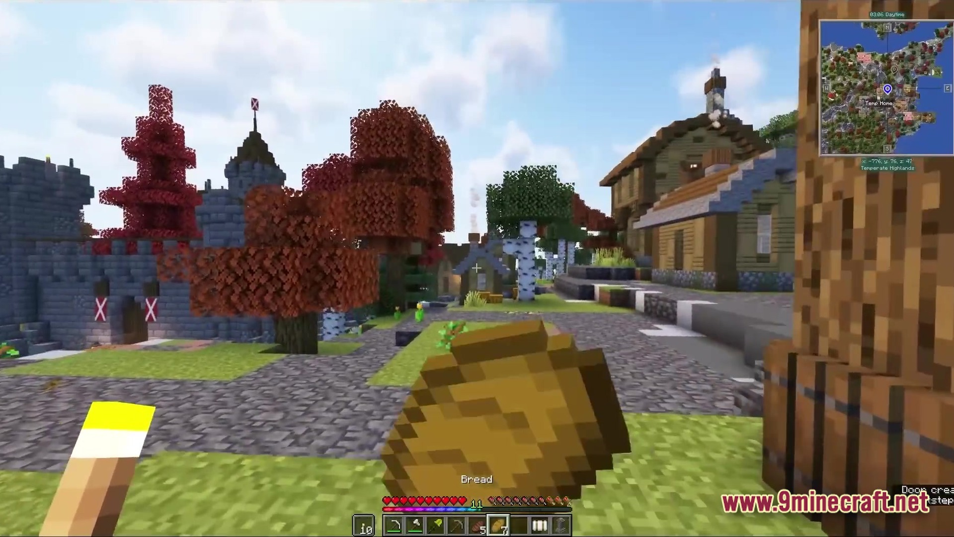 A player holds bread while standing in a village with custom houses, trees, and a stone castle