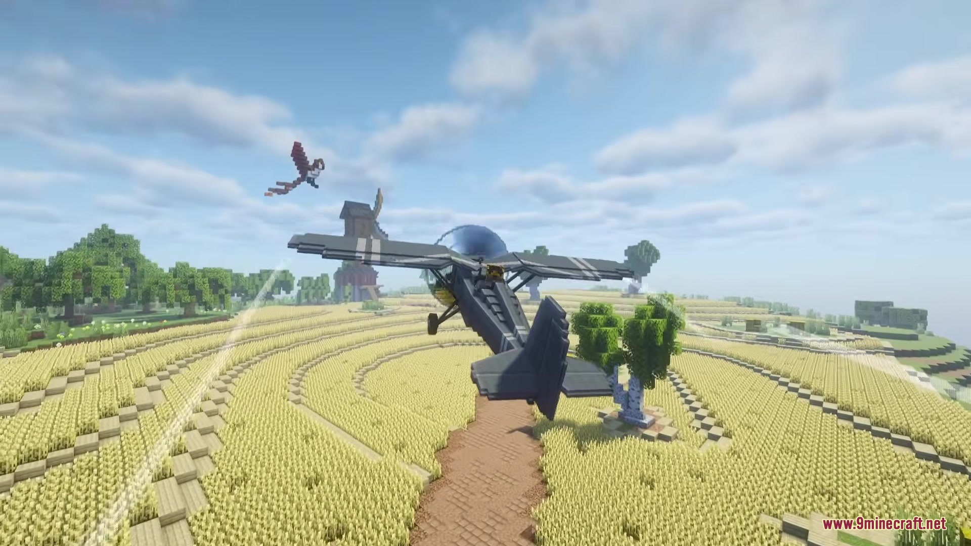 A grey civilian plane flies over a golden wheat field with a windmill