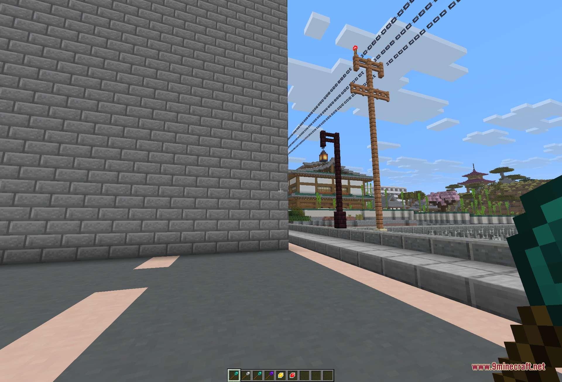 A tall grey stone-brick wall raised in one action beside a paved road, with the wand and its crafting materials sitting in the hotbar and a Japanese-style town in the background