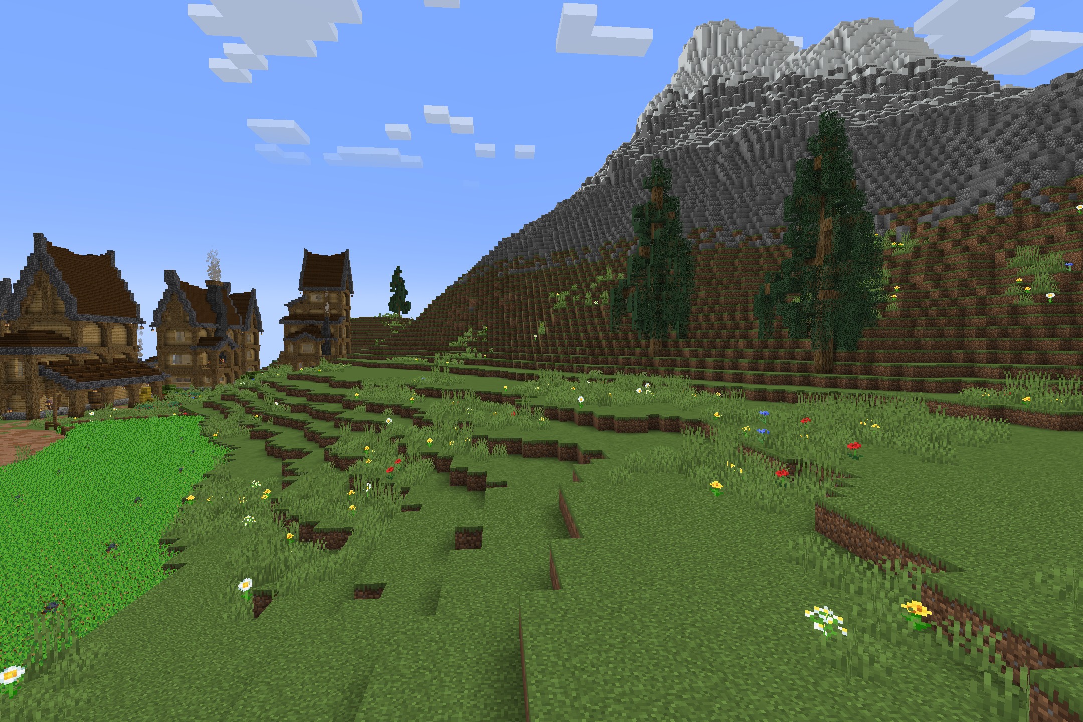 Grassy hills with flowers and spruce trees next to a rocky mountain