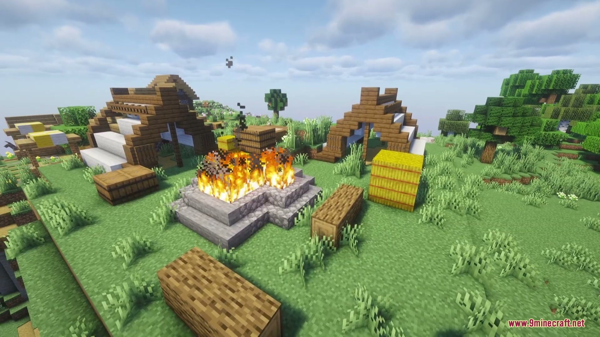 A large campfire burns in a historical camp with wooden structures and hay